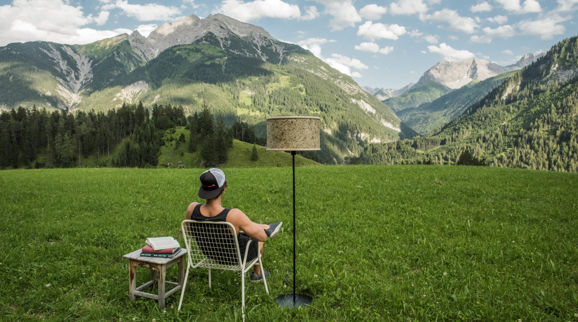 Ulrich Huber sitzt auf einem Stuhl und genießt die Aussicht auf die Alpen neben ihm steht ein Nachttisch und eine Lampe von ALMUT von Wildheim