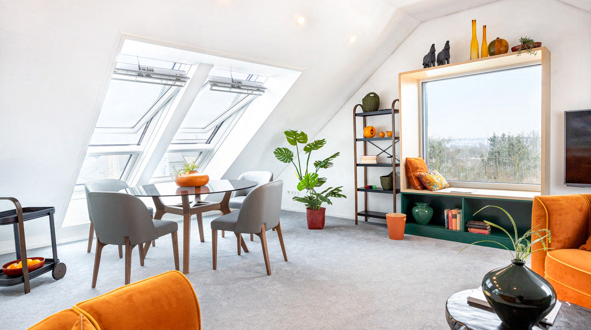Das Dachgeschoss mit schöner Aussicht ins Wuppertal. Es ist hell gestaltet und wirkt dadurch trotz Dachschrägen groß. Man sieht einen Teppichboden, Stühle samt Tisch, Deko, Pflanzen, Sessel, normales und Dachfenster.